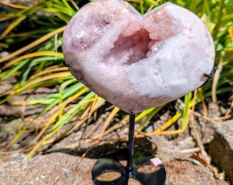 May include: A heart-shaped amethyst geode with light purple and white crystalline textures, mounted on a black metal stand with a circular base. The geode features a central hollow cavity filled with small, sparkling purple crystals.