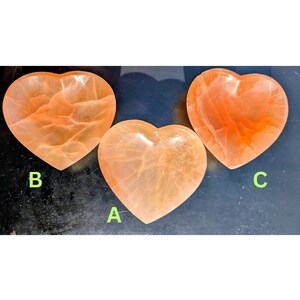 May include: Three heart-shaped, orange-colored crystal bowls. Each bowl has a unique pattern and texture. The bowls are labeled A, B, and C. The bowls are displayed on a dark surface.