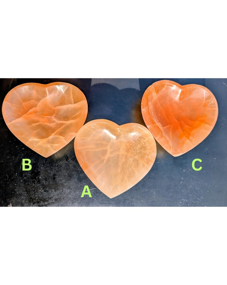May include: Three heart-shaped, orange-colored crystal bowls. Each bowl has a unique pattern and texture. The bowls are labeled A, B, and C. The bowls are displayed on a dark surface.