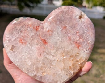 May include: A heart-shaped crystal stone with a mix of pink, white, and orange hues. The stone has a rough, textured surface and is held in a hand. The background is blurred, showing greenery and a sunny outdoor setting.