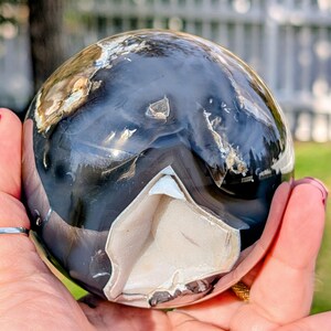 May include: A polished agate sphere with a mix of black, white, and tan colors. The sphere has a smooth, reflective surface with a unique pattern. The sphere is held in a hand, showcasing its size and detail.