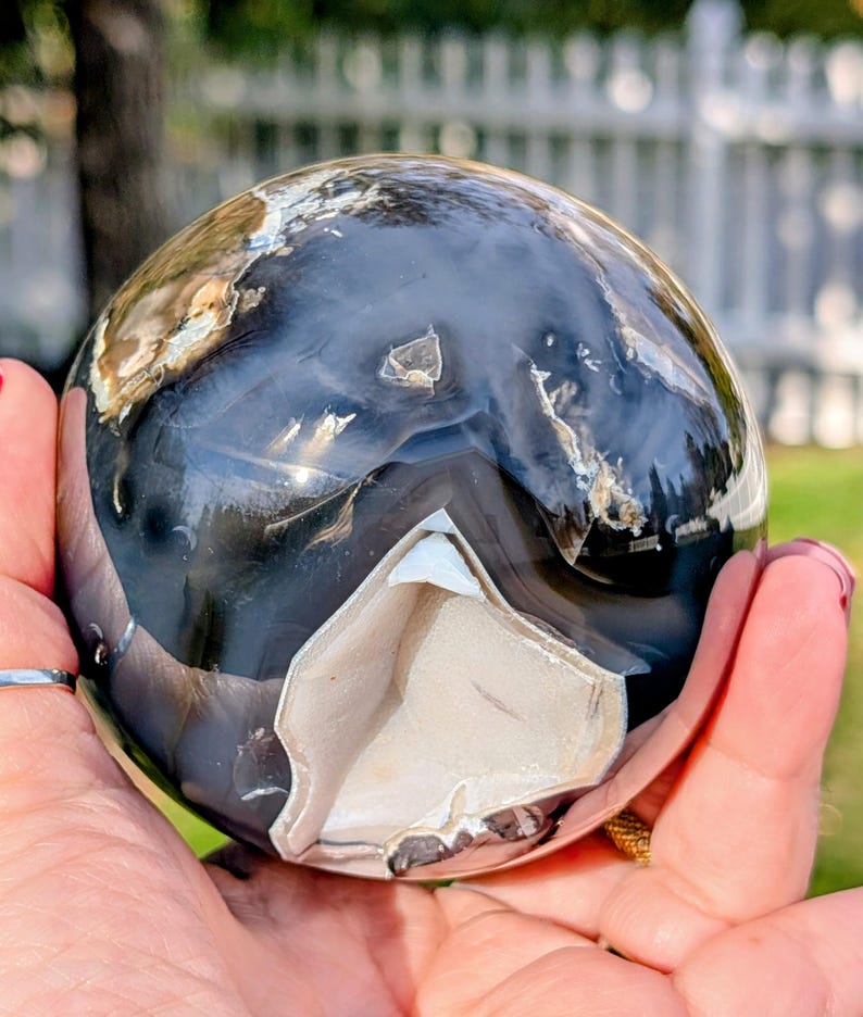 May include: A polished agate sphere with a mix of black, white, and tan colors. The sphere has a smooth, reflective surface with a unique pattern. The sphere is held in a hand, showcasing its size and detail.