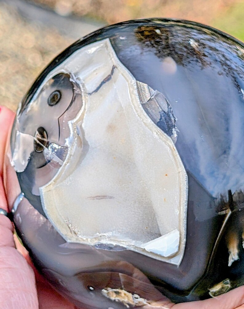 May include: A polished, spherical gemstone with a striking pattern. The stone features a mix of dark gray, black, and creamy white colors. The surface has a glossy finish, and the unique veining creates an eye-catching design. The gemstone is held in a hand.