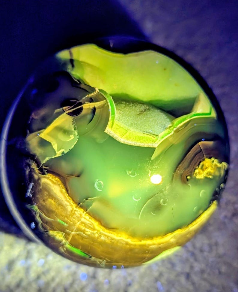 May include: A close-up of a clear, round glass marble with a swirling pattern of green, yellow, and brown colors. The marble has a polished surface and a unique, abstract design. The colors are vibrant and create a sense of depth.