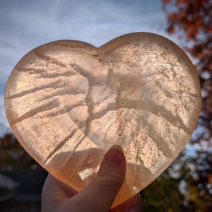 May include: A translucent, heart-shaped stone with a warm, peach hue. The stone has natural veining patterns and is held up against a backdrop of a blue sky and autumn foliage. The heart is a symbol of love and affection.