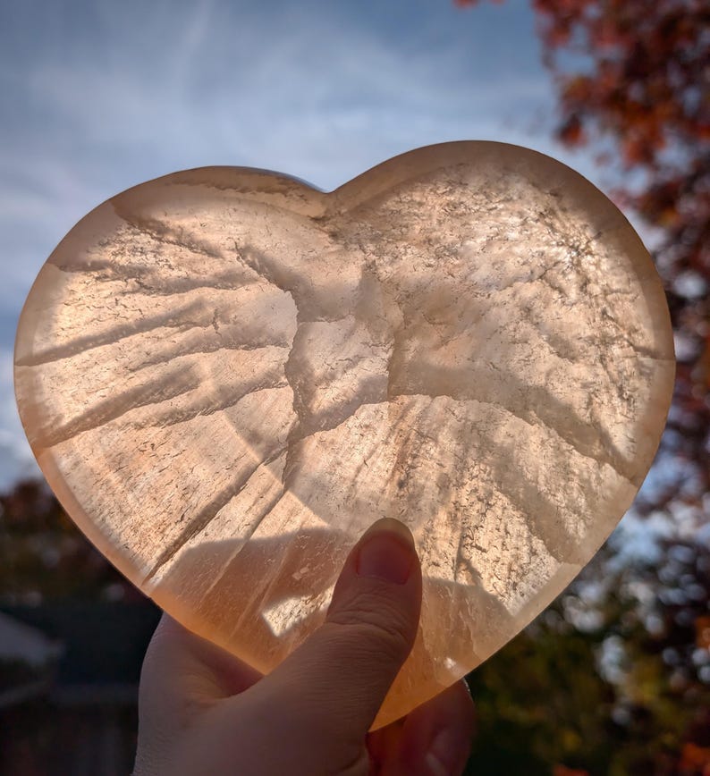 May include: A translucent, heart-shaped stone with a warm, peach hue. The stone has natural veining patterns and is held up against a backdrop of a blue sky and autumn foliage. The heart is a symbol of love and affection.