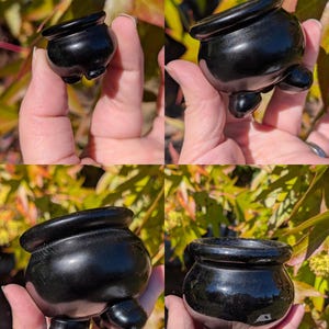 Pu&ograve; includere: Un piccolo oggetto nero lucido a forma di calderone, con tre piedini arrotondati e un bordo svasato. Il piccolo vaso ha una superficie liscia e riflettente, probabilmente realizzata in pietra lucida o materiale simile. L'oggetto &egrave; tenuto in una mano.