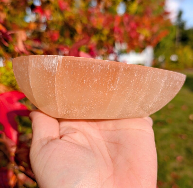 May include: A peach-colored, oval-shaped bowl carved from a translucent stone. The bowl is held in a hand, with the background showing fall foliage. The bowl's surface has visible striations, adding to its natural aesthetic.