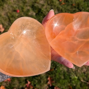 May include: Two heart-shaped, translucent orange-colored crystal bowls. The bowls are held in a hand, with the sunlight reflecting off the smooth surfaces. The bowls are made of a natural stone material.