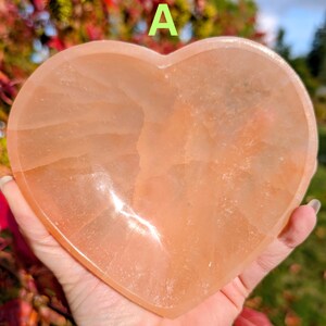 May include: A heart-shaped, peach-colored crystal. The stone is held in a hand, with the background showing colorful foliage and the letter "A". The crystal has a smooth, polished surface.