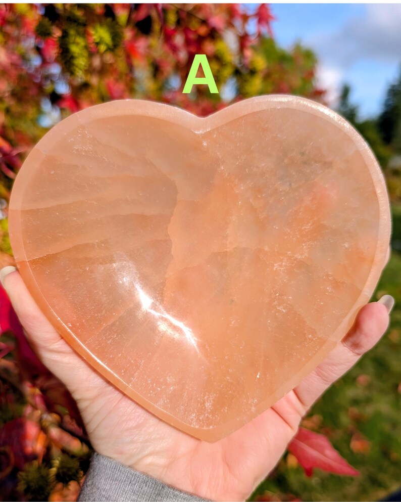 May include: A heart-shaped, peach-colored crystal. The stone is held in a hand, with the background showing colorful foliage and the letter "A". The crystal has a smooth, polished surface.