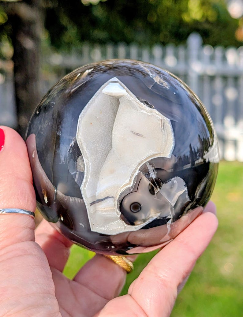 May include: A polished, spherical geode with a striking contrast of black and white patterns. The geode's interior reveals a crystalline structure, creating a unique and eye-catching decorative object. The sphere is held in a hand, showcasing its size and detail.