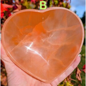 May include: A heart-shaped, peach-colored crystal bowl. The bowl is held in a hand and has a smooth, polished surface. The letter "B" is visible in the background. The bowl is likely used for decorative purposes or as a small dish.