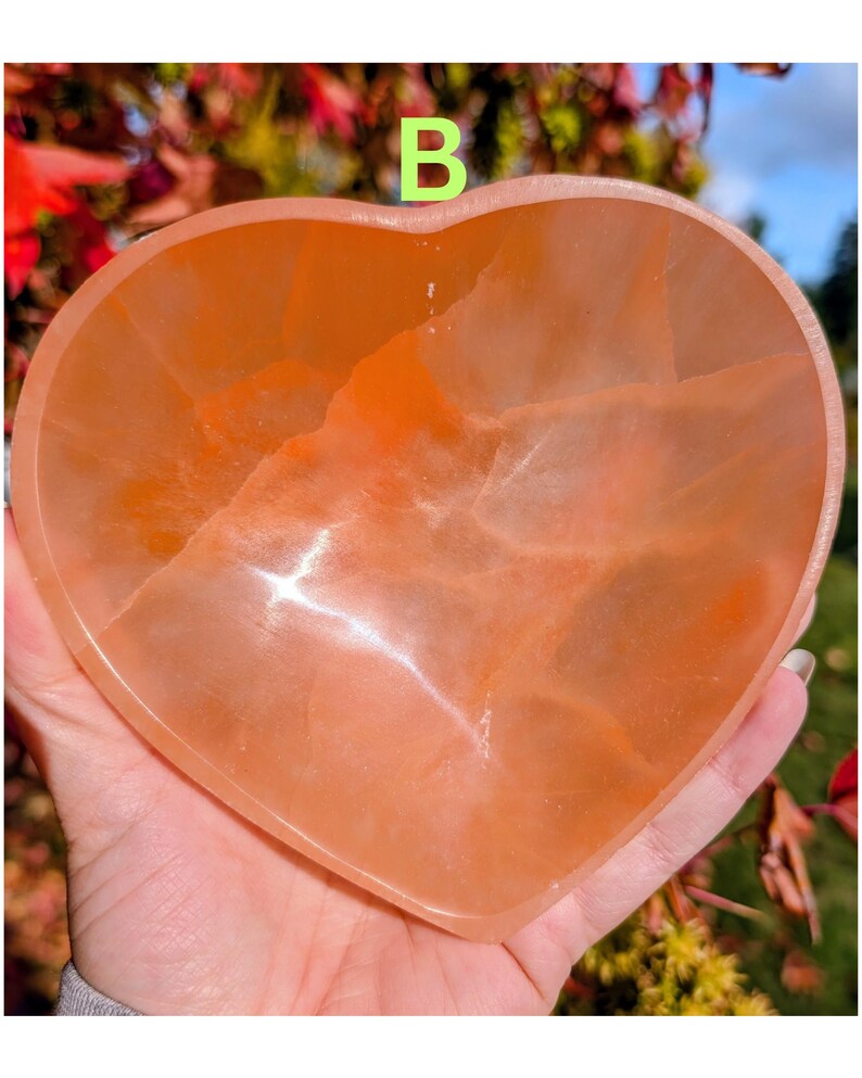 May include: A heart-shaped, peach-colored crystal bowl. The bowl is held in a hand and has a smooth, polished surface. The letter "B" is visible in the background. The bowl is likely used for decorative purposes or as a small dish.