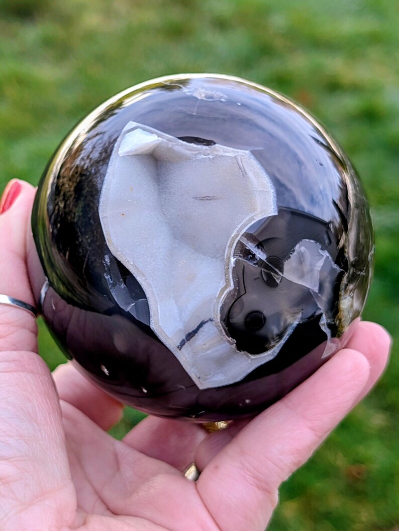 May include: A polished black and white agate sphere. The sphere has a geode-like cavity with white crystalline formations. The surface reflects light, and the sphere is held in a hand against a blurred green background.