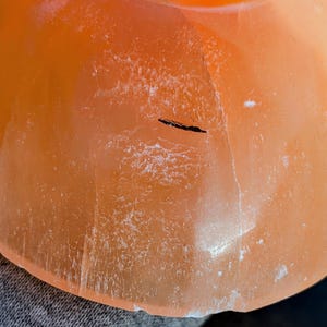 May include: Close-up of a translucent orange-colored object with a rounded shape. The surface has a textured appearance with white crystalline deposits and a dark linear inclusion. The object is likely a decorative item or a piece of natural stone.