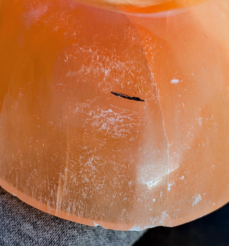 May include: Close-up of a translucent orange-colored object with a rounded shape. The surface has a textured appearance with white crystalline deposits and a dark linear inclusion. The object is likely a decorative item or a piece of natural stone.