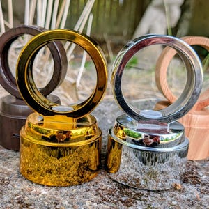 May include: A collection of circular display stands featuring a reflective gold metallic base and a reflective silver metallic base, each topped with a matching circular ring frame. Additional wooden-textured circular stands are visible behind.