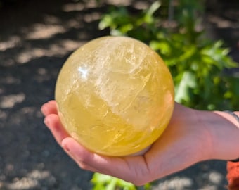 May include: A translucent, yellow crystal sphere is held in an open hand. The sphere has a smooth, rounded shape and a slightly textured surface. The background is blurred, with green foliage and a gravel path visible.