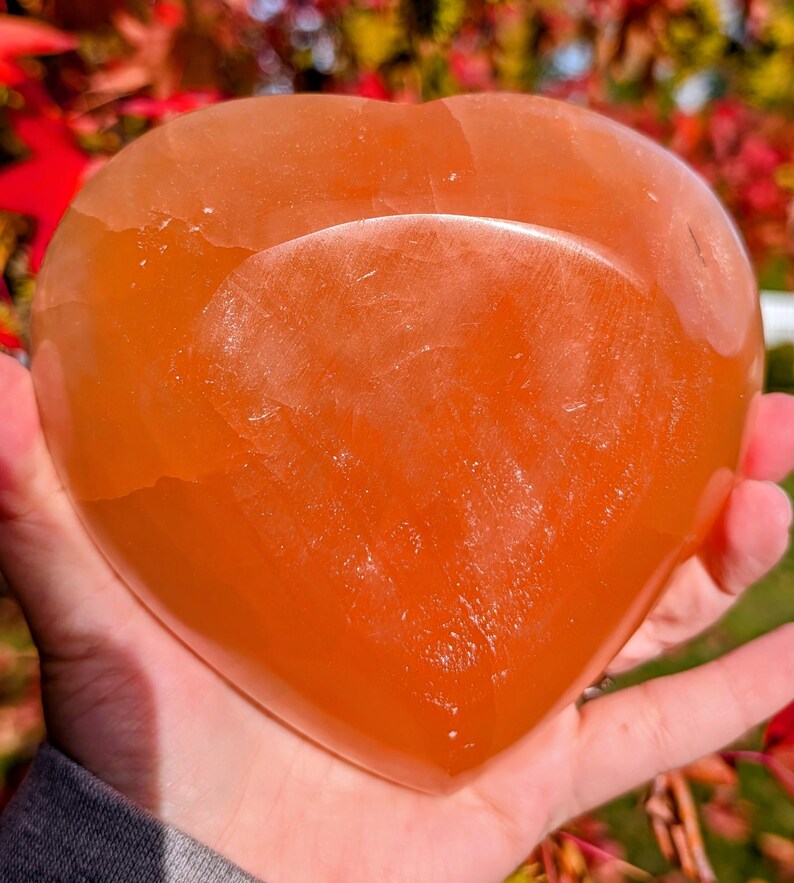 May include: A heart-shaped orange crystal stone. The stone is smooth and polished, with a translucent appearance. The crystal is held in a hand, with a blurred background of red and green foliage. The heart shape is clearly defined.