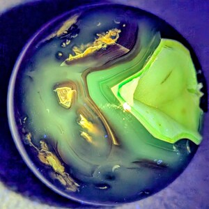 May include: A close-up view of a polished stone with a swirling pattern of green, gold, and blue hues. The stone has a layered appearance, with bands of color creating a unique, abstract design. The overall effect is reminiscent of a miniature landscape.