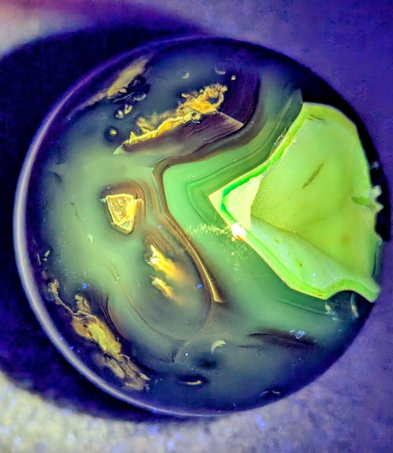 May include: A close-up view of a polished stone with a swirling pattern of green, gold, and blue hues. The stone has a layered appearance, with bands of color creating a unique, abstract design. The overall effect is reminiscent of a miniature landscape.