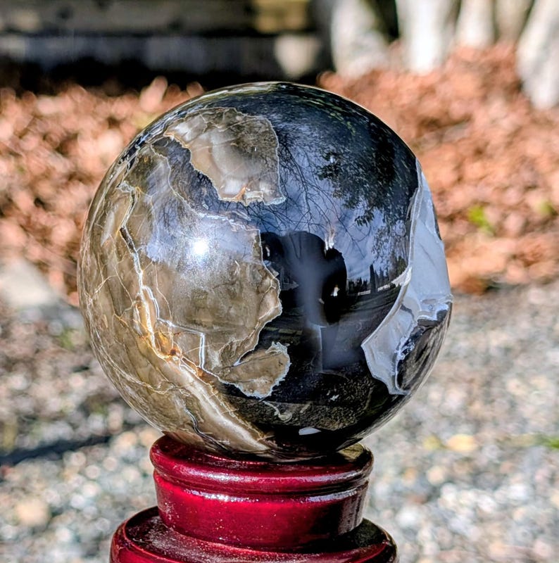 May include: A polished, spherical stone with a mix of dark and light brown, gray, and black colors. The sphere sits on a red wooden stand. The surface of the sphere reflects the surrounding environment.