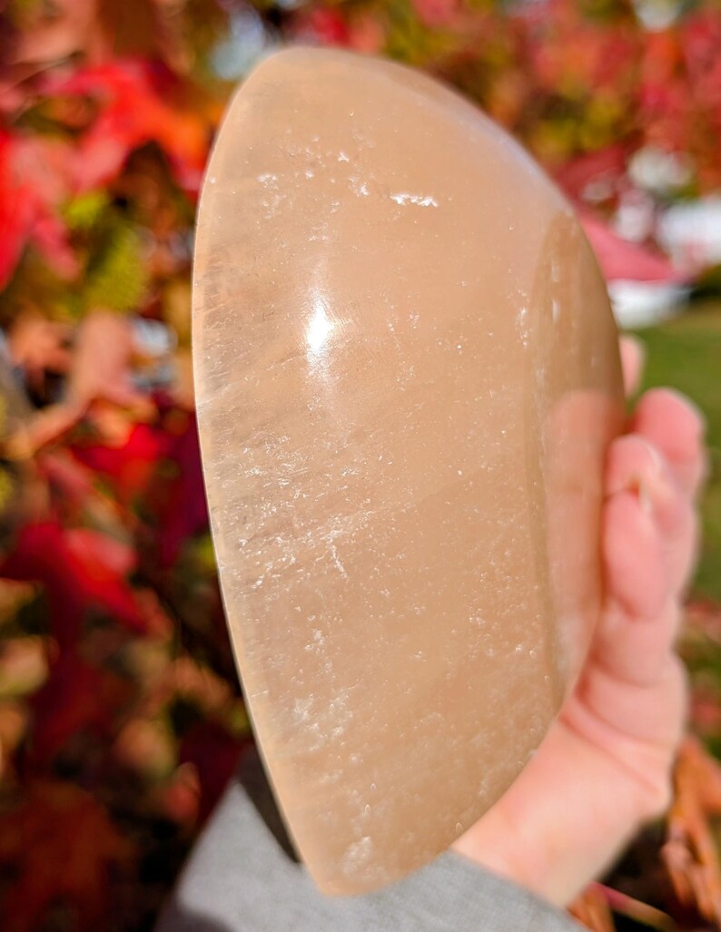 May include: A smooth, peach-colored stone with a rounded shape. The polished surface reflects light, highlighting subtle variations in color and texture. The stone is held in a hand, with a blurred background of fall foliage.