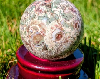 May include: A decorative sphere with a unique pattern of green, brown, and cream colors, resting on a glossy, deep red stand. The sphere's surface has a natural, organic appearance, and the stand is a rich, contrasting color.