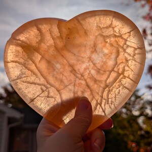 May include: A translucent, heart-shaped stone with a warm, peachy-orange hue. The stone has intricate internal veining, creating a unique pattern. The heart is held up against a backdrop of a cloudy sky and autumn foliage.