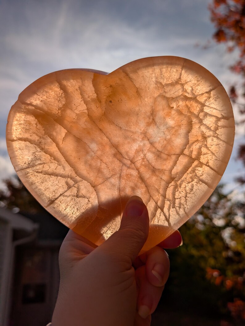 May include: A translucent, heart-shaped stone with a warm, peachy-orange hue. The stone has intricate internal veining, creating a unique pattern. The heart is held up against a backdrop of a cloudy sky and autumn foliage.