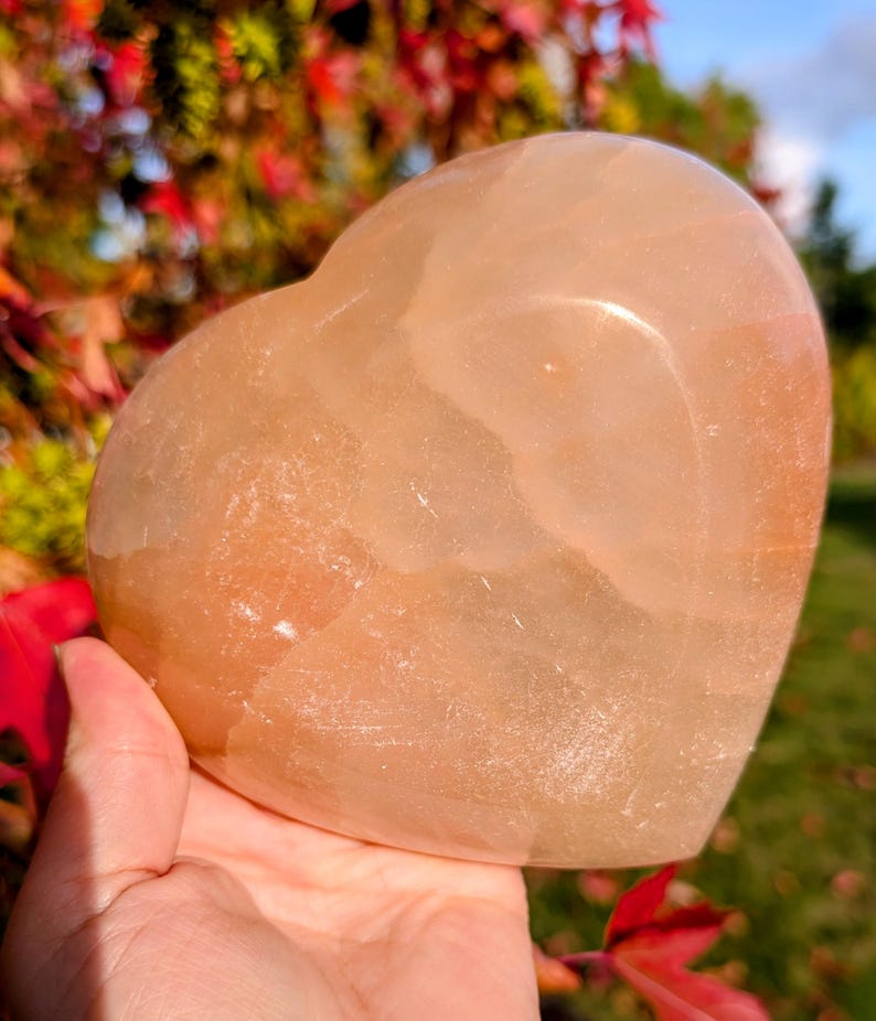 May include: A peach-colored heart-shaped stone, held in a hand. The stone has a smooth, polished surface with subtle variations in color. The background features colorful autumn foliage.