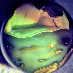 May include: A close-up of a translucent, spherical object with layers of green, yellow, and brown hues. The object has a smooth, polished appearance with internal structures visible. The lighting creates a glowing effect, highlighting the colors and textures.
