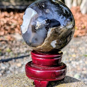 May include: A polished sphere of petrified wood, displaying a mix of dark and light brown tones with white inclusions. The sphere sits atop a glossy, deep red wooden stand. The stand is composed of three stacked, circular tiers.
