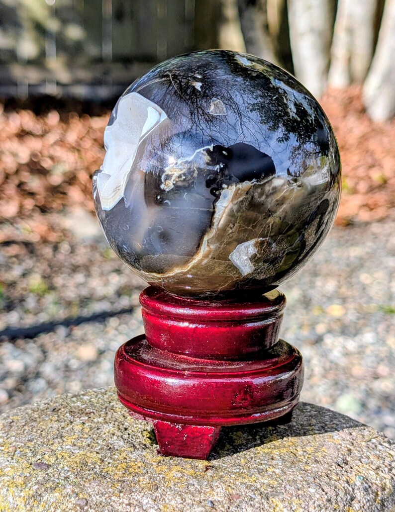 May include: A polished sphere of petrified wood, displaying a mix of dark and light brown tones with white inclusions. The sphere sits atop a glossy, deep red wooden stand. The stand is composed of three stacked, circular tiers.