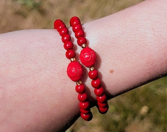 May include: Two beaded bracelets featuring small, round, vibrant red beads interspersed with tiny gold-toned spacers and a single, larger, oval-shaped red bead with a textured, embossed pattern centred on each strand.