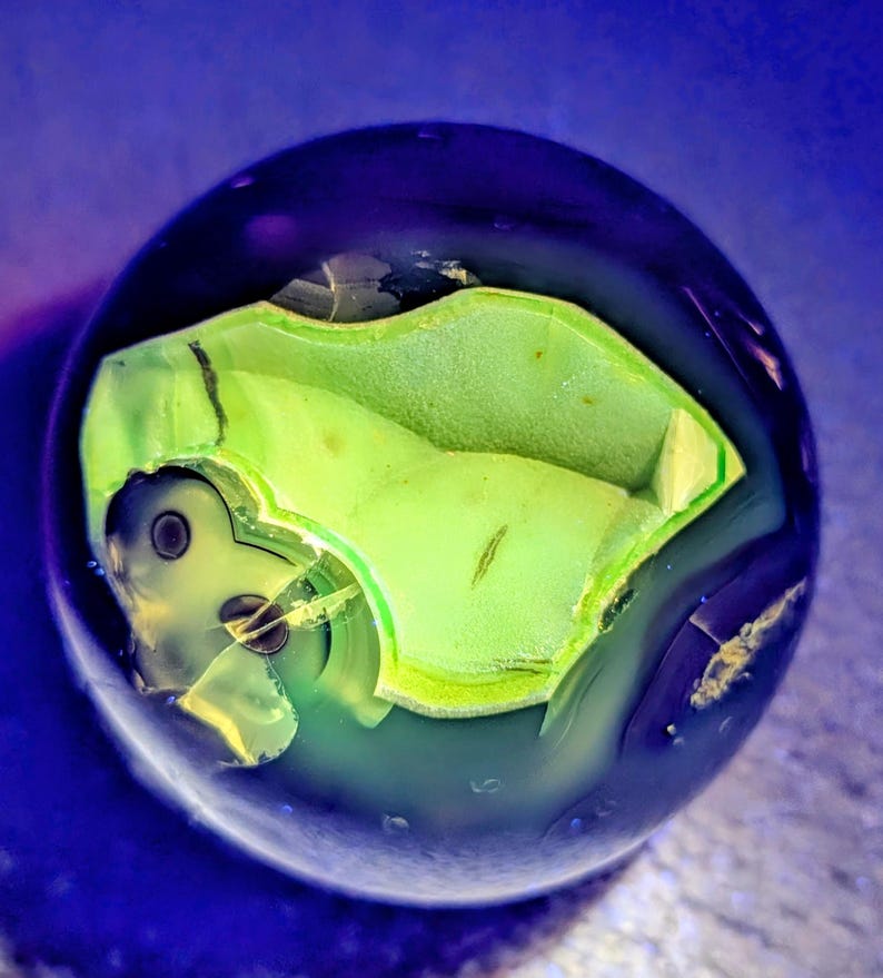May include: A spherical, polished gemstone with a dark blue exterior and a vibrant green interior. The interior features a unique, irregular shape with a lighter green hue, creating a striking contrast. The gemstone's surface reflects light, highlighting its smooth texture.