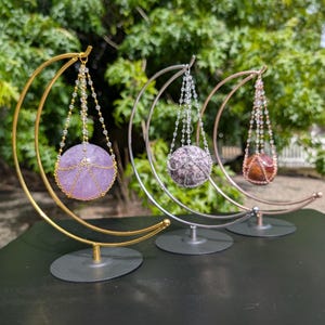 May include: Three decorative sphere holders with crescent moon-shaped frames. The spheres are purple, gray, and orange, suspended by beaded chains. The frames are gold, silver, and rose gold, each on a circular base. The background is blurred greenery.