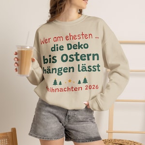 Könnte beinhalten: Ein beigefarbener Rundhals-Pullover mit dem deutschen Text 'Wer am ehesten ... die Deko bis Ostern hängen lässt' in Rot und Dunkelgrün, ergänzt durch kleine grüne Tannenbaum-Symbole und den roten Schriftzug 'Weihnachten 2026' am unteren Rand.
