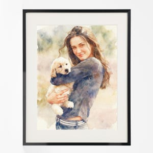 Könnte beinhalten: Aquarellporträt einer Frau, die einen Golden Retriever-Welpen hält. Das Kunstwerk ist in einem schwarzen Rahmen gerahmt. Die Frau trägt ein dunkelblaues Hemd und Jeans. Der Hintergrund hat weiche Aquarellwaschungen in Grün, Gelb und Rosa.