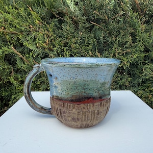 May include: Handmade ceramic mug with a blue, green, and brown color scheme. The top of the mug is light blue, transitioning to green and brown. The bottom has a textured, brown, basket-weave design. The mug has a curved handle.