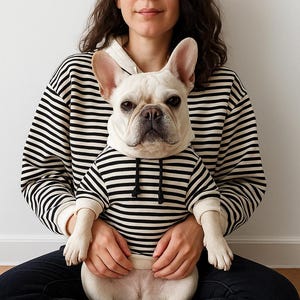 Puede incluir: Una persona y un bulldog francés blanco con sudaderas a rayas blancas y negras a juego. El perro está sentado en el regazo de la persona. La persona lleva una sudadera a rayas blancas y negras y vaqueros azul oscuro.