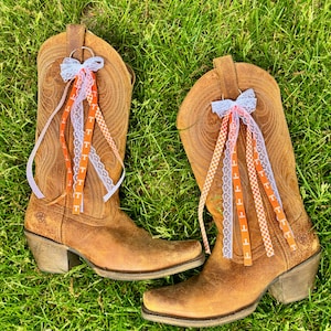 Op de afbeelding: Een paar bruine leren cowboylaarzen met decoratieve linten. De linten zijn oranje en wit, met de letter "T" erop gedrukt. De laarzen staan op groen gras.