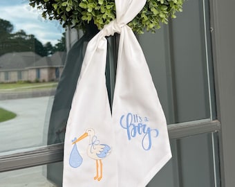 May include: A white fabric door hanger with a green wreath. The hanger features a stork carrying a blue bundle and the text "It's a Boy" in blue script. The design is perfect for a baby shower or welcoming a newborn.