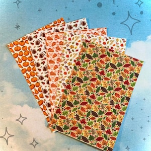 Puede incluir: Una colección de hojas de papel estampadas con diseños de temática otoñal. Los patrones incluyen calabazas, porciones de tarta, castañas y coloridas hojas de otoño en tonos naranja, marrón y verde. Las hojas están dispuestas en forma de abanico sobre un fondo azul.