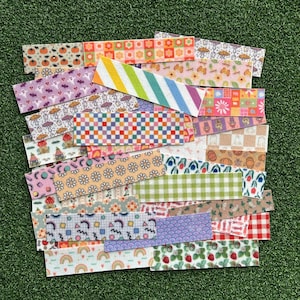 Op de afbeelding: Een verzameling decoratieve stoffen stroken met verschillende patronen, waaronder pompoenen, geesten, paddenstoelen, regenbogen en bloemmotieven. De stroken hebben verschillende kleuren, zoals oranje, paars, groen en roze, en zijn gerangschikt op een groene ondergrond.