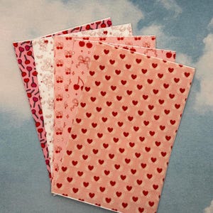 Könnte beinhalten: Ein Fächer aus fünf rosa- und weiß gemusterten Papieren. Die Muster zeigen rote Herzen, Kirschen, Lollis und Engel. Die Papiere sind vor einem bewölkten blauen Hintergrund angeordnet.