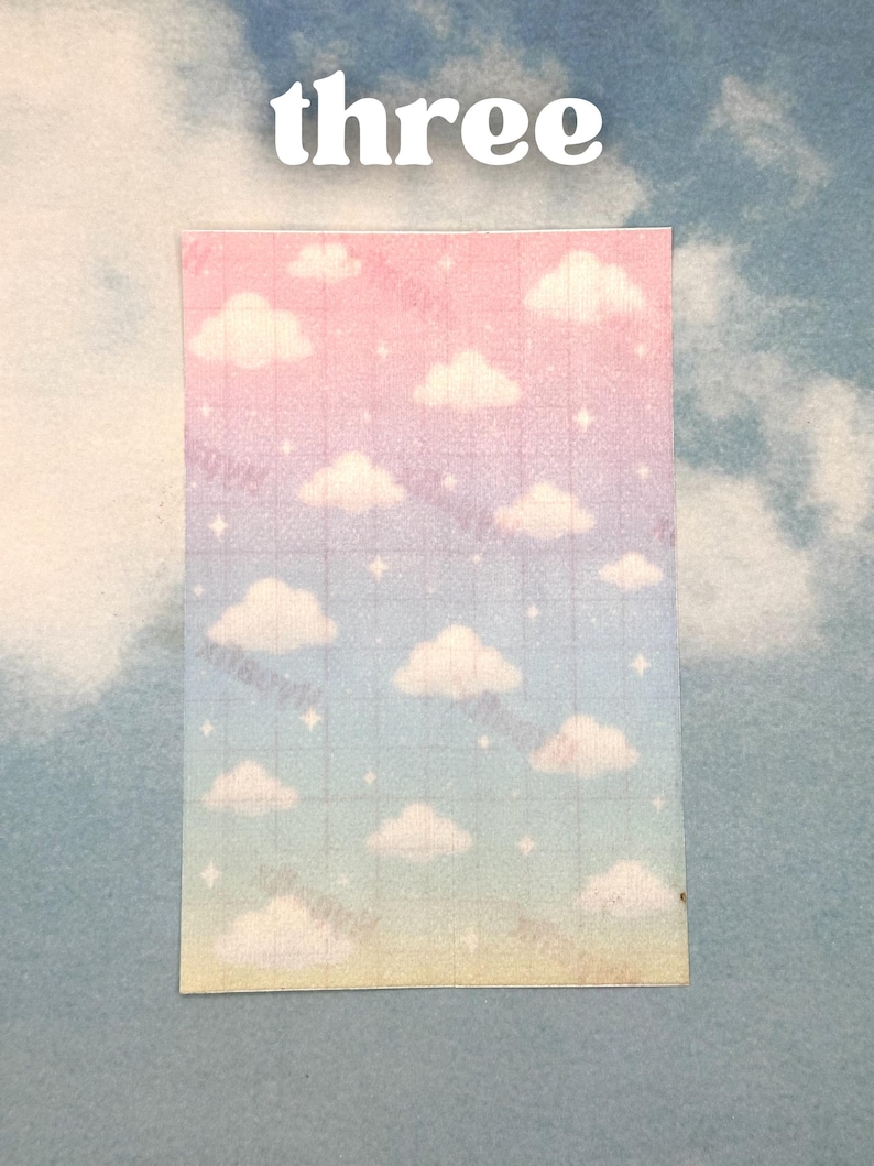 Puede incluir: Una pegatina rectangular con un dise&ntilde;o de cielo degradado, con nubes y estrellas. La pegatina tiene un patr&oacute;n de cuadr&iacute;cula y la palabra "three" en letras blancas en la parte superior. El fondo es un cielo azul claro lleno de nubes.