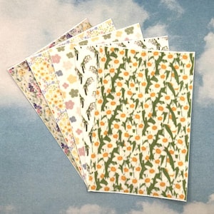 Op de afbeelding: Een verzameling papieren met bloemmotieven, uitgespreid tegen een hemelsblauwe achtergrond. De papieren hebben verschillende ontwerpen, waaronder madeliefjes, lavendel en andere kleurrijke bloemen. De papieren zijn rechthoekig en lijken bedoeld te zijn voor knutselen of scrapbooking.