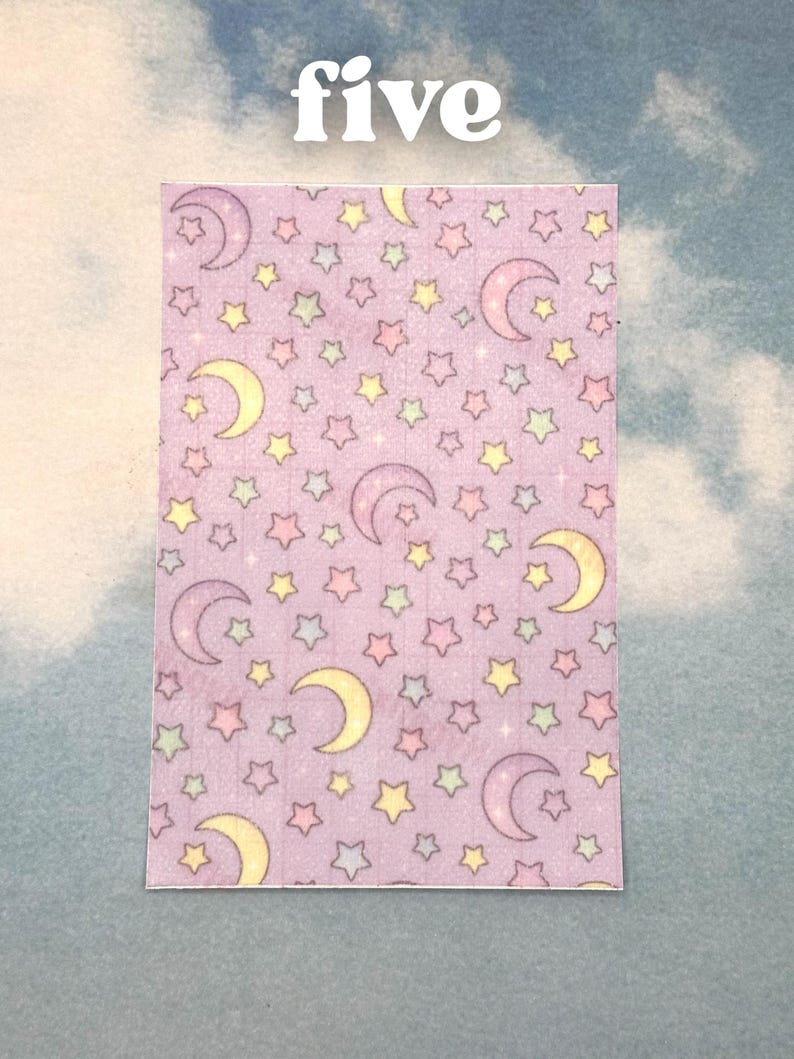 Puede incluir: Un trozo de papel rectangular con un patr&oacute;n repetitivo de estrellas amarillas y lunas crecientes en tonos de morado, rosa y verde sobre un fondo lavanda. La palabra "five" est&aacute; escrita en blanco en la parte superior.